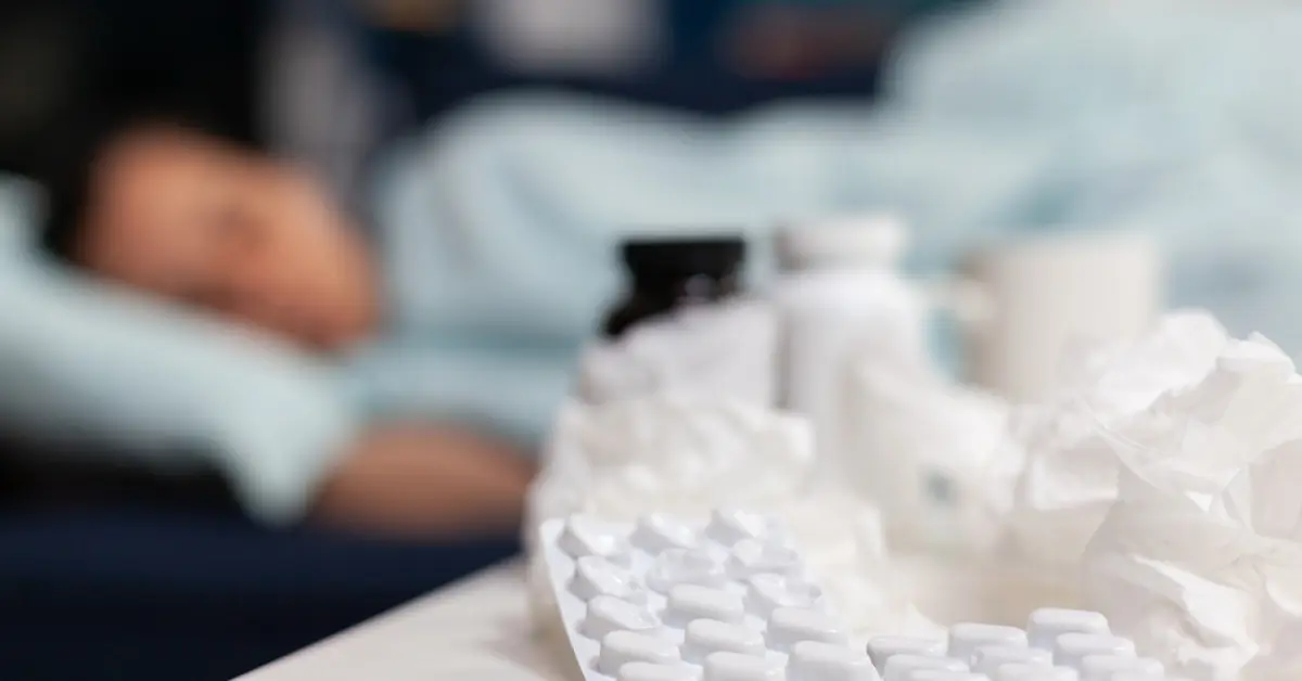 A blurred figure resting on a bed with a teal blanket, surrounded by medication, tissues, and a cup on a table